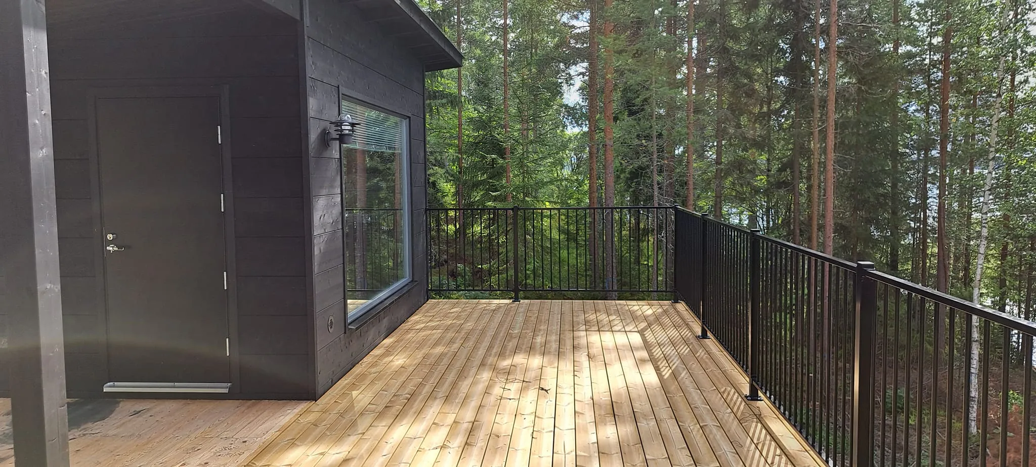 Musta talo puuterassilla, metsä taustalla. Aurinkoinen sää.