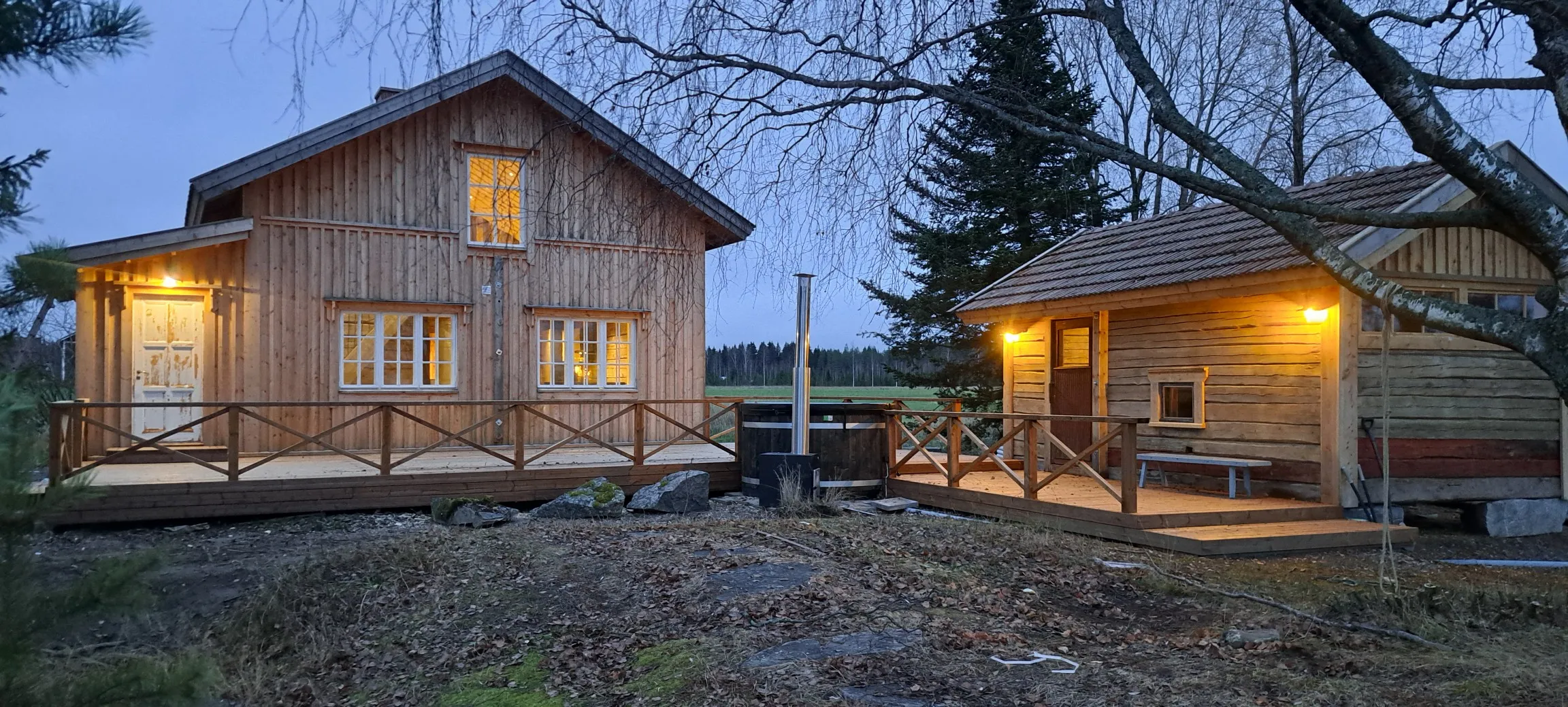 Kaksi puurakenteista taloa, joissa on valaistut ikkunat ja terassit, sekä puinen kylpytynnyri ulkona hämärässä maisemassa.